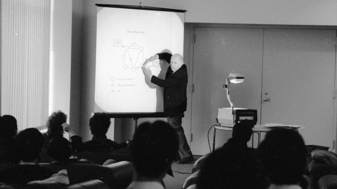 Black-and-white image of Mischa Schwarz gesturing toward a projected diagram while teaching in a classroom.