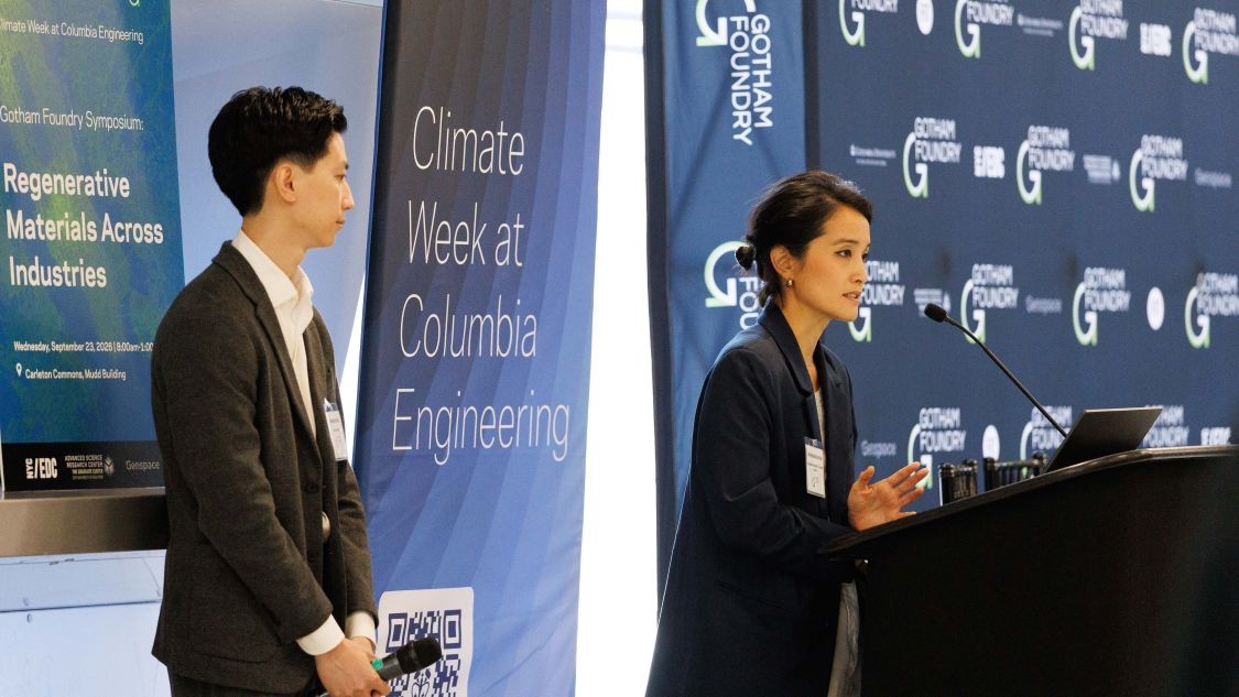 Shiho Kawashima speaking from a podium while Diandian Zhao stands to the side looking toward the speaker at the Gotham Foundry symposium.
