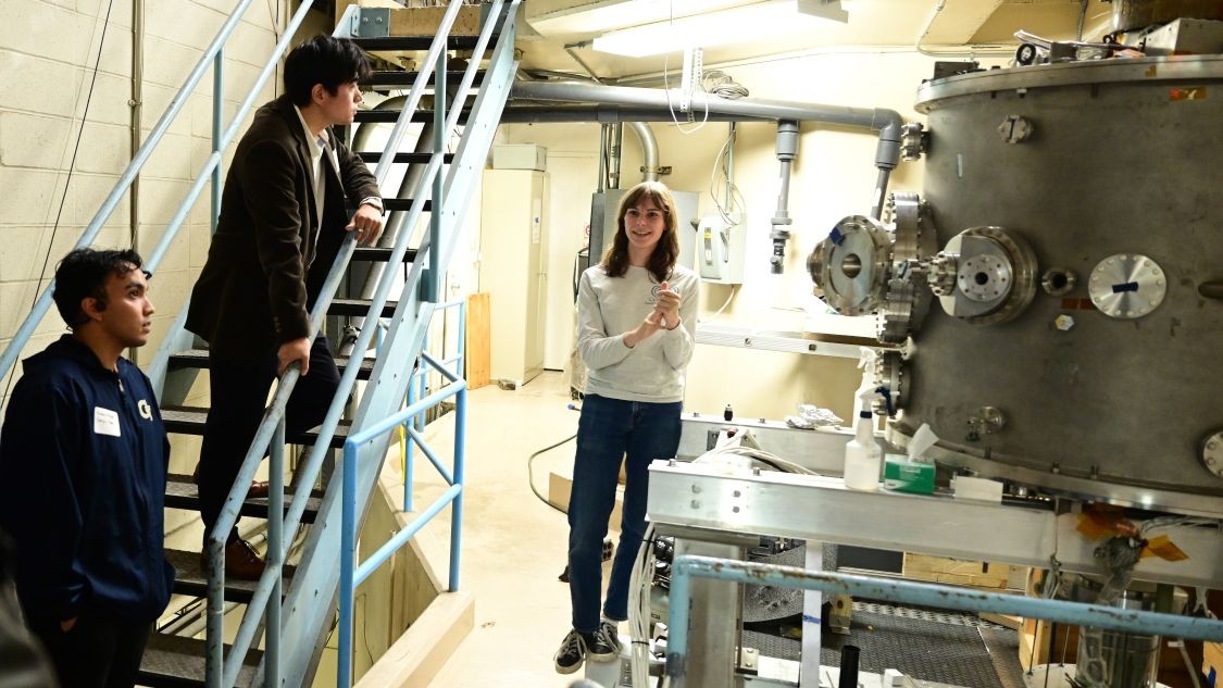 Three people stand beside fusion research equipment during a lab tour, with two attendees observing from a staircase.