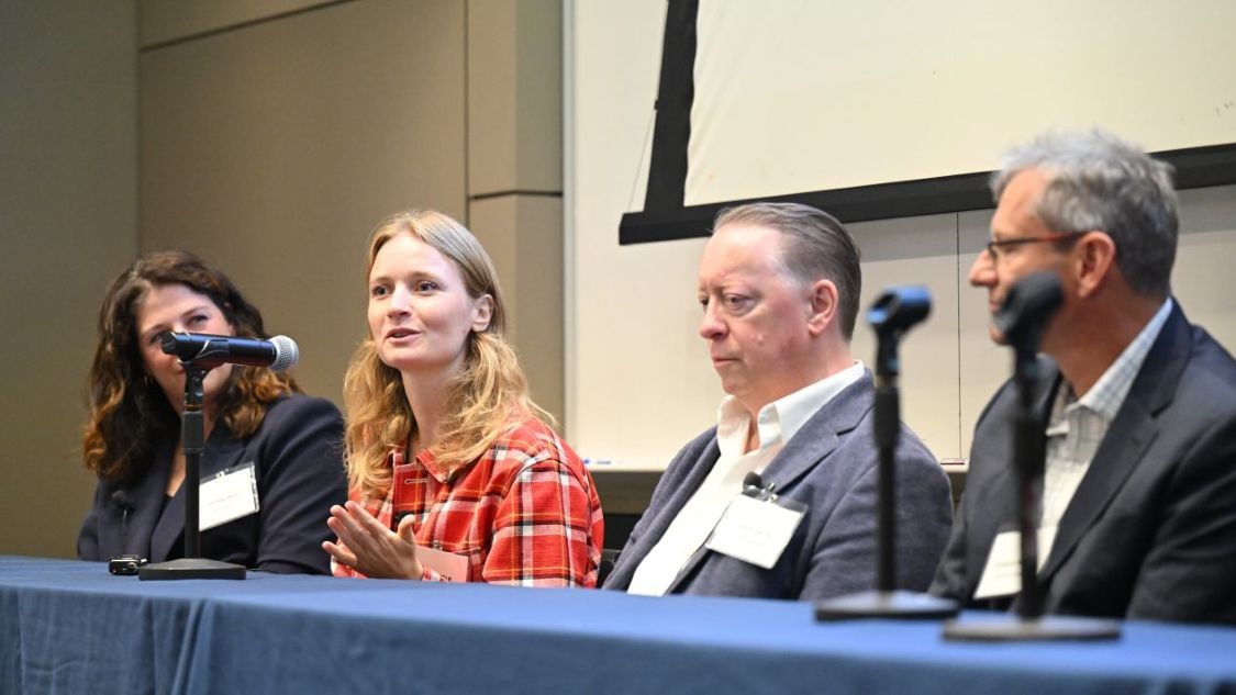 Four panelists sit at a table during a panel discussion, with one speaking and gesturing.