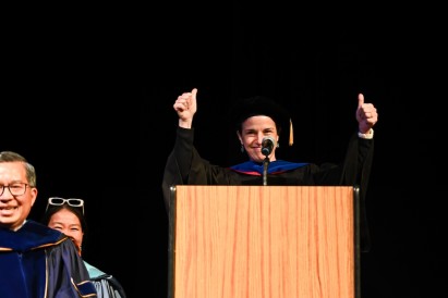 Elisa Konofagou in graduation regalia holding two thumbs up behind a podium with a microphone