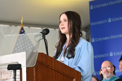 Columbia Engineering valedictorian Danielle Maydan in a light blue graduation gown speaks into a microphone at a wooden podium inside a tent, with a Columbia University backdrop and an American flag behind her.