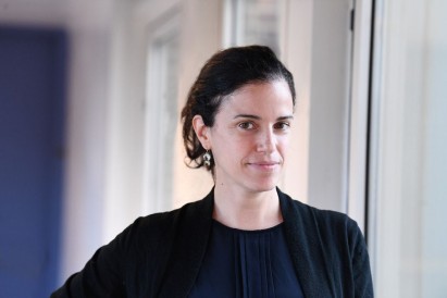 Potrait photo of Elisa E. Konofagou dressed in a black jacket over a dark blue shirt, looking into the camera in a well-lit hallway with white walls and a blue door.