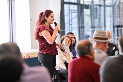Sandra Soueid speaks into a microphone while holding a notebook, addressing seated attendees during a roundtable discussion in a sunlit room.